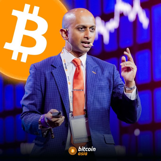 A bald speaker in a blue checkered suit and orange tie presents at Bitcoin Asia, gesturing while holding a remote, with a large Bitcoin logo and stock chart backdrop.