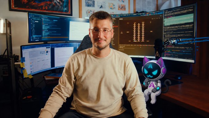 A man with glasses and a beige sweater sitting at a desk with multiple monitors displaying code and data, accompanied by a cartoon robot cat graphic.
