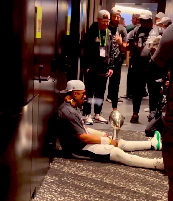 A player in a white cap and dark jersey sits on the floor in a hallway, smoking a cigar next to the Vince Lombardi Trophy (Super Bowl trophy) while teammates in celebratory attire stand behind him.