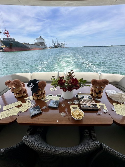 A view from a boat shows a wooden table covered with teddy bears wearing merchandise, flowers, snacks, and scattered flyers, with large ships visible in the harbor behind.