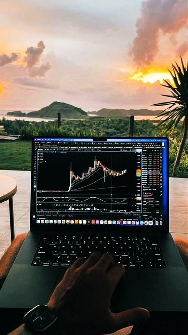 A person using a MacBook laptop showing a trading chart while overlooking a scenic sunset view of mountains and ocean.