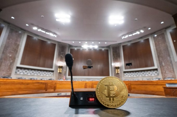 A physical Bitcoin coin sits prominently on a congressional hearing room desk beside a microphone, symbolizing the crypto regulatory debate at the heart of the CLARITY Act, which aims to clarify whether the SEC or CFTC will oversee digital assets. The empty chamber underscores the urgency of the bill's narrow window to pass, as lawmakers work to finalize crypto jurisdiction rules.