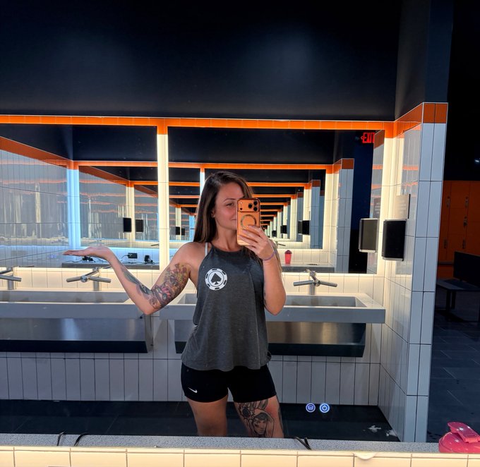 A woman takes a mirror selfie in a gym locker room while wearing a gray tank top with a logo, paired with black Nike shorts. Her toned arm and visible thigh tattoos highlight her fitness dedication, prompting her playful question about whether it's time to "burn" the shirt as part of her transformation journey.
