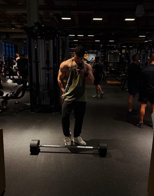 A fit man in a green tank top takes a mirror selfie in a gym, posing confidently with a trap barbell at his feet as other gym-goers work out in the background—capturing a classic "GM" (good morning) post showcasing his gym dedication and physique.