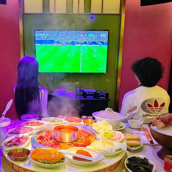 Two people sit at a table loaded with hot pot dishes and side ingredients, watching a soccer match on a wall-mounted TV showing an "ETV" broadcast of a game on a vibrant green field. The setup captures the social moment of enjoying a meal while following a match, fitting the caption's mention of watching their home team's game and reacting to DR Congo scoring first.