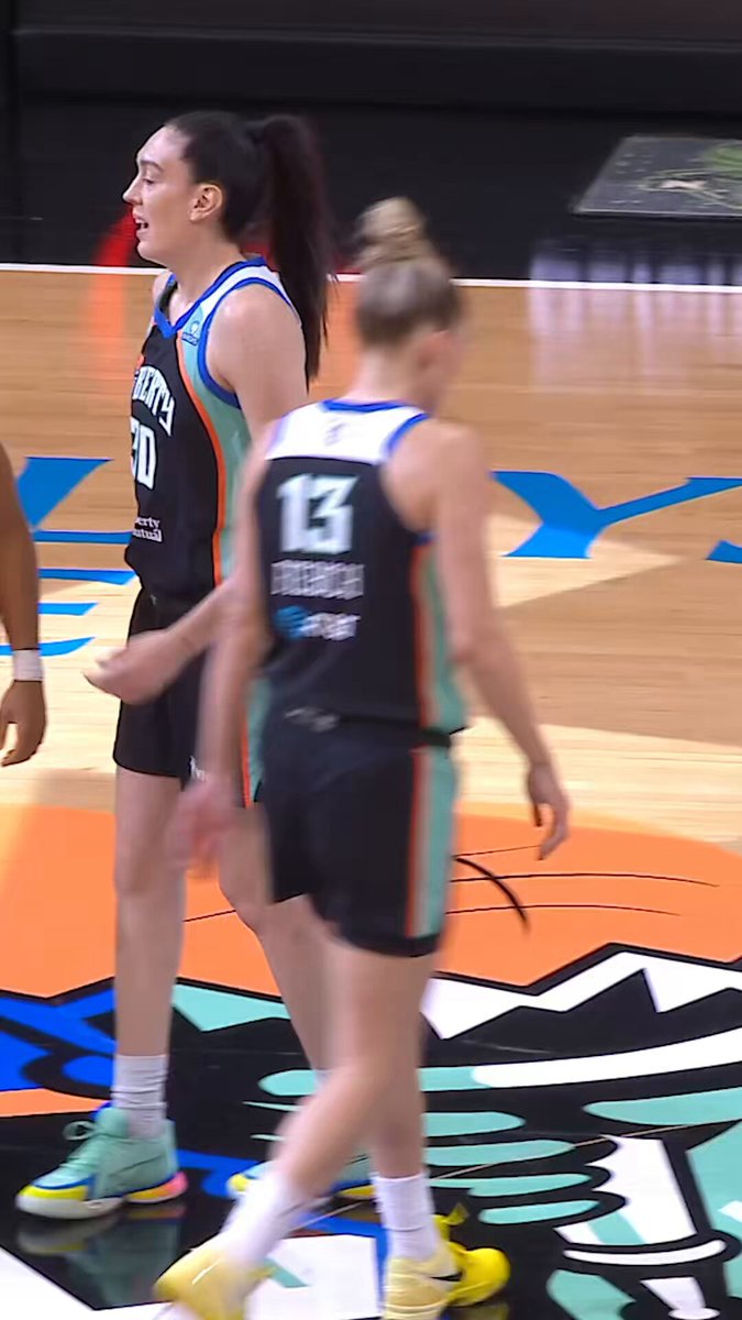 The image shows two female basketball players standing on a basketball court, one of them wearing a black jersey with the number 13 while the other has a blue jersey. They are both smiling as they look at the camera. The background reveals that this is an indoor sports facility, and there's a sign in the background that reads "Volume Up @ coinbase brings you inside the game with Mic'd Up Moments." This suggests that the image captures a moment during a basketball game where the players are enjoying themselves and sharing their experience with others.