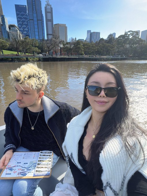 A selfie of a young couple sitting on a boat, with the woman wearing sunglasses and a white cardigan, set against a backdrop of a river with city skyscrapers in the distance.