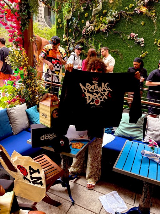 A group of people at a lifestyle or brand event, with one person holding up a black shirt with 'Rituals of a Doer' text. A box labeled 'designed dog' and 'JOBS' are also visible.
