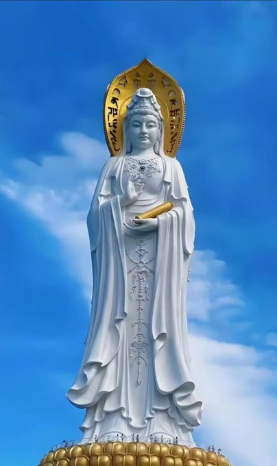A tall white statue of Guanyin (Goddess of Mercy) with a golden halo and sacred markings, standing against a blue sky.