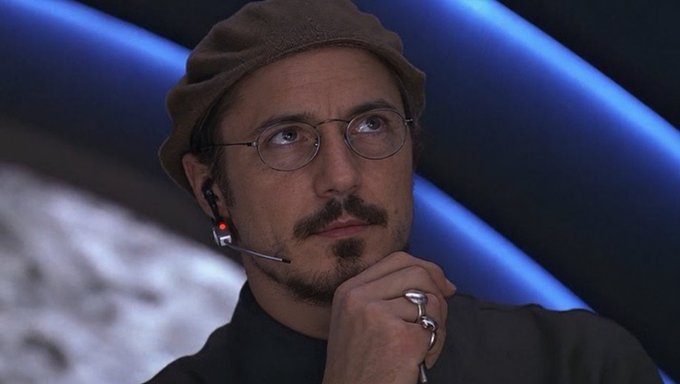 Photograph of a young man wearing a brown beanie, round wire-frame glasses, with a goatee and mustache, wearing an earpiece and silver rings, in a contemplative pose.
