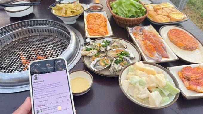A spread of raw meats, seafood, and vegetables is arranged on a table around a charcoal grill, with a smartphone in the foreground displaying a crypto price-tracking app.