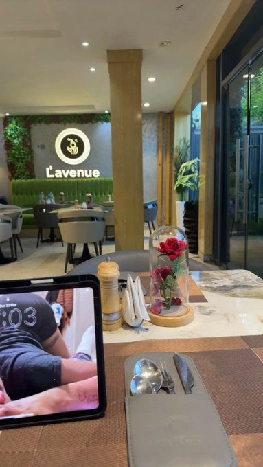 A tablet displaying the time "5:03" rests on a restaurant table at "L'avenue," alongside a rose under a glass cloche, cutlery in a leather pouch, and a salt shaker, while the caption humorously motivates followers for an early Monday morning routine.