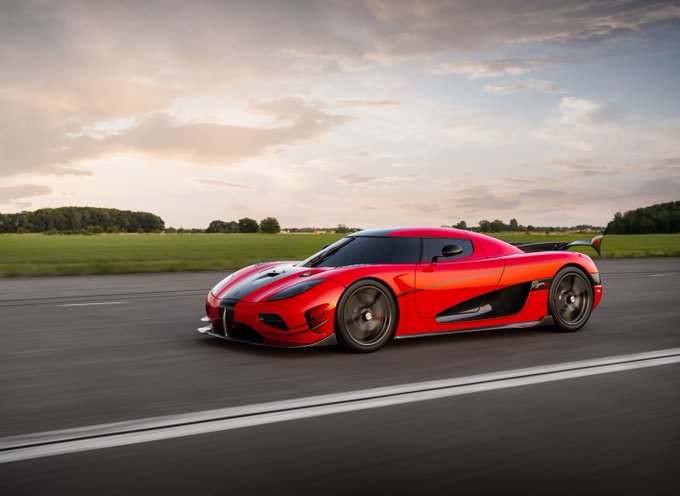 A red Koenigsegg Agera sports car driving on a paved road with a sunset sky and green fields in the background.