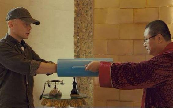 A scene showing two men exchanging a blue folder, with one wearing a black cap and jacket, and the other in a red traditional robe, set in a tiled room.
