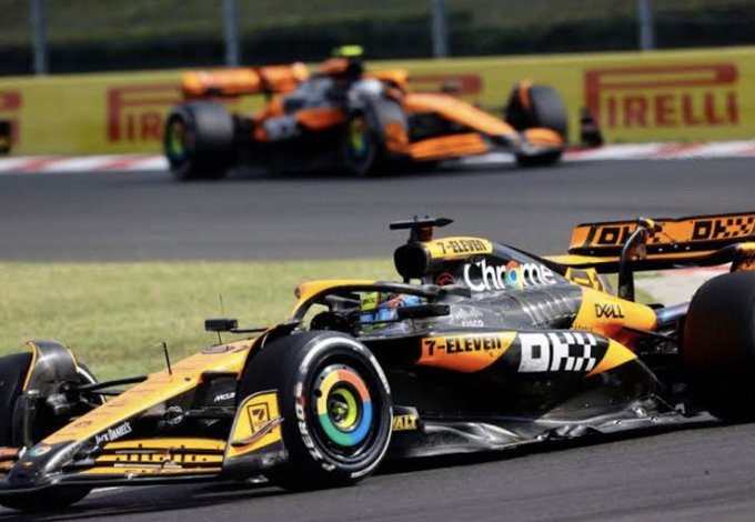 Two Formula 1 race cars on a track, with a McLaren car prominently in the foreground featuring 'Chrome' and 'DHX' sponsorships, and an orange McLaren car visible in the background near a Pirelli banner.