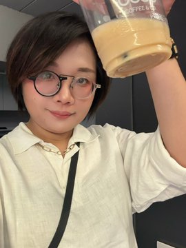 A person with short dark hair and round glasses wearing a light-colored shirt, holding up an iced coffee drink.