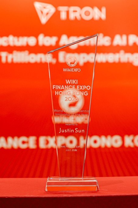 A glass trophy presented to Justin Sun at the Wiki Finance Expo Hong Kong in July 2026, where he was named "World's Most Influential Innovator of the Year," displayed in front of a TRON-branded orange backdrop. The award aligns with the post's message of appreciation to WikiExpo for the recognition and TRON's continued collaboration with BAI.