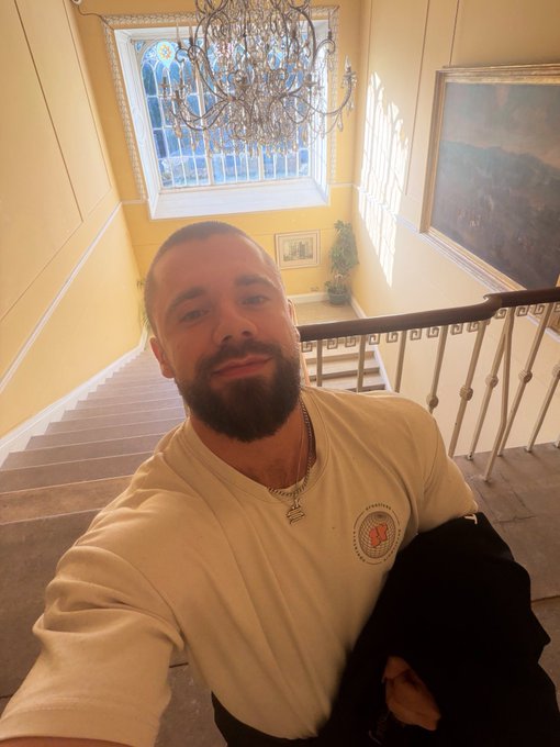 A bearded man takes a selfie at the top of an elegant staircase in a building with yellow walls, a large crystal chandelier, and stained glass windows.