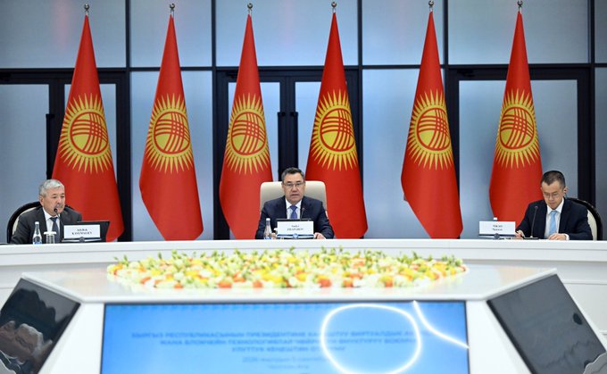 Officials sit at a conference table during a diplomatic meeting, with five Kyrgyzstan flags displayed prominently in the background behind a floral centerpiece.