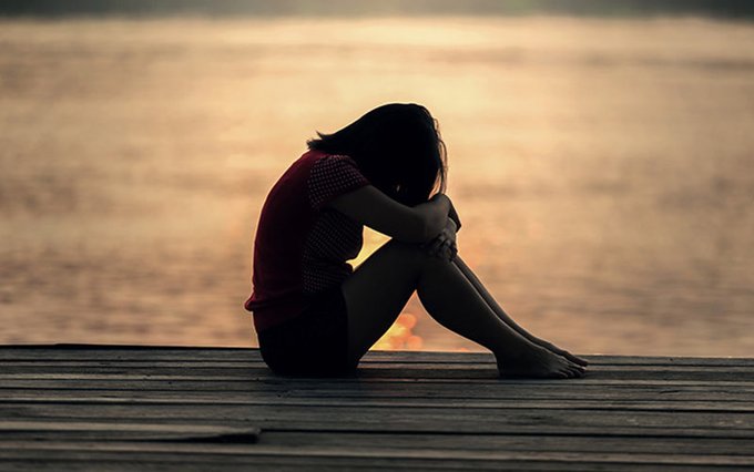 A silhouette of a woman sitting on a wooden dock at sunset, hugging her knees with her head down, conveying a melancholic mood.