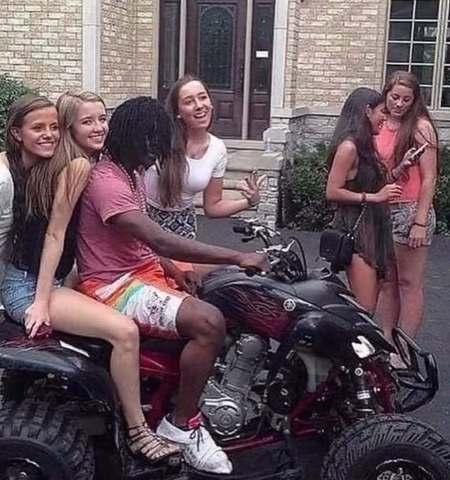 A group of six smiling teenage girls gather around a black and red Yamaha motorcycle, with two seated on the bike and the others standing close by, posing cheerfully in front of a brick house.