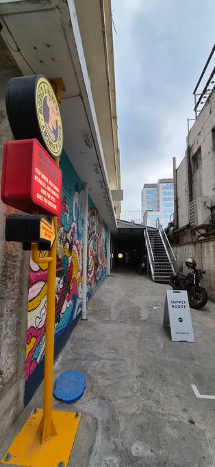Street photography shot of an alleyway with colorful graffiti murals, a yellow and red signal post in the foreground, and a 'SUPPLY ROUTE' sign visible near a staircase.