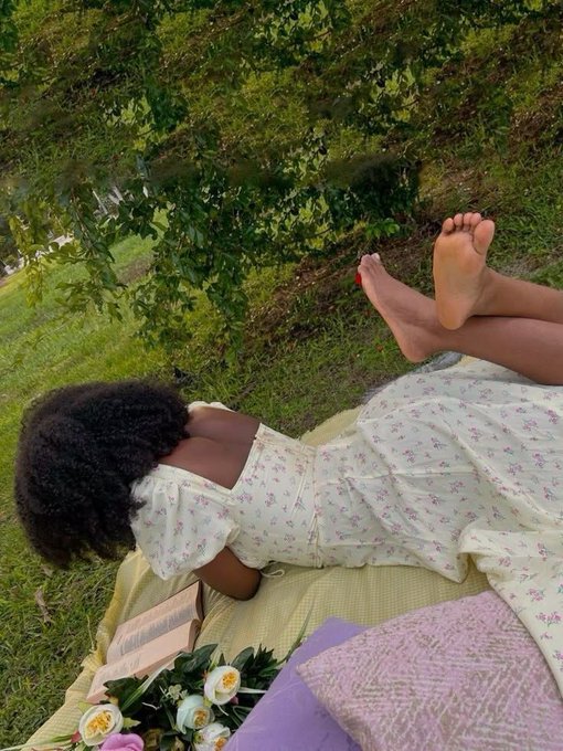 A woman with curly hair relaxes on a yellow blanket in a grassy outdoor setting, wearing a floral sundress with her bare feet propped up and a bouquet of white and pink flowers nearby, capturing a serene weekend picnic vibe.
