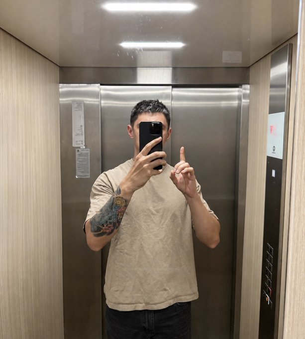 A man takes a mirror selfie in an elevator, holding up his index finger in a gesture that echoes the "one trade" message of the post—symbolizing the single opportunity needed to change everything. The sleek metallic elevator interior and the tattoo on his forearm complete the confident, motivational vibe of the "gm" greeting.