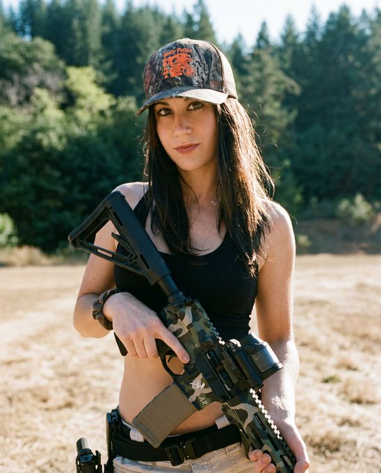 A young woman stands outdoors in a grassy field holding a camouflage AR-15 style rifle, wearing a black tank top and a camo 'Trump 2020' cap.