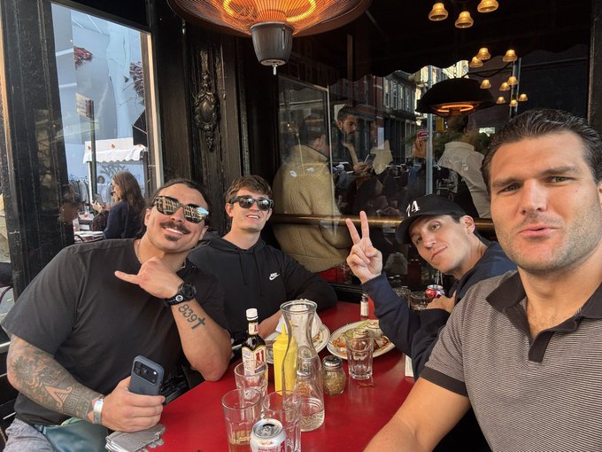 The image shows a group of four men sitting at an outdoor restaurant table, posing for a photo with their arms around each other's shoulders. They appear to be enjoying themselves as they share a meal together. The table is set with various items such as cups, bottles, and utensils like forks and knives. There are also chairs placed around the dining table. In the background, there are additional people sitting at tables or standing near them, indicating that this outdoor restaurant is quite popular.