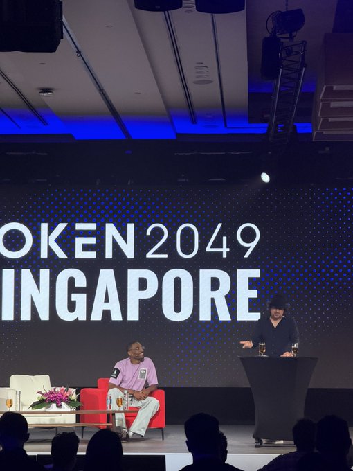 A panel at "Token 2049 Singapore" features two men on stage—one seated in a red chair in a purple shirt, the other standing at a podium in a black shirt—likely with the man in purple being the focus of the post's anticipation of being "roasted," while the post expresses bullish sentiment on the ticker $ANSEM.