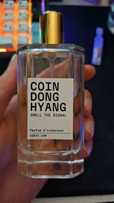 A hand holds a clear glass perfume bottle with a gold cap and a white label reading "COIN DONG HYANG SMELL THE SIGNAL, Parfum d'intérieur, upbit.com" in front of a backlit mechanical keyboard.