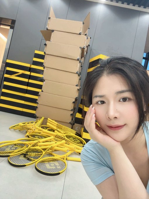 A woman poses for a selfie in front of stacks of yellow "Binance" branded cardboard boxes and yellow pickleball paddles, with the post celebrating Binance's 9th anniversary and her 5.8 years with the platform. The branded merchandise in the background ties directly to her message of appreciation for Binance.