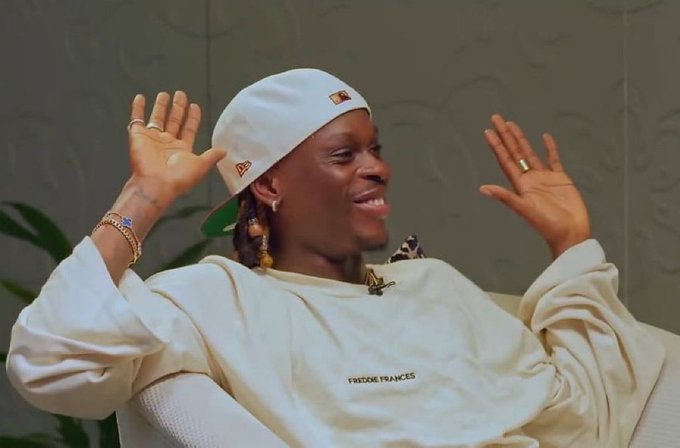 The image features a man wearing a white hat, sitting down with his arms raised up in the air while smiling at the camera. The background of the photo shows a wall that has a plant behind him, adding some greenery to the scene. There is also an object on the floor near the bottom right corner of the image.

The post appears to be about a Zora fashion brand and their coin collection. It seems like the man in the white hat might have made money from selling coins or promoting the brand through his social media presence, as indicated by the caption "Hi mutuals How was your day? Did you make any money today? Well I made some from my Zora profile coin Let me know how your day was in the CS".