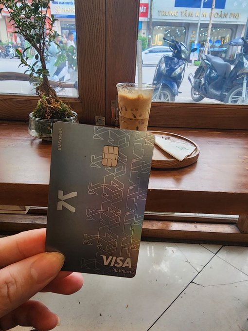 A hand holds up a KAST Visa Platinum business card in the foreground, with an iced coffee and a small bonsai-like plant on a wooden windowsill behind it, looking out onto a street with parked motorbikes and a sign reading "TRUNG TÂM THỜI TRANG FN."

The image showcases the physical KAST Visa Platinum card in a real-world setting—a café—visually reinforcing the post's message about KAST making digital and stablecoin-based finance tangible and accessible in everyday life.