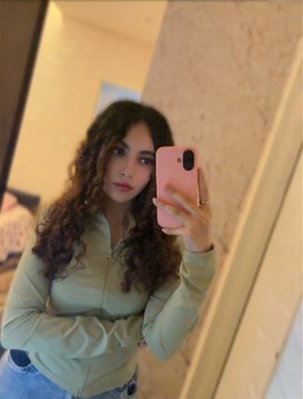 A young woman with long curly brown hair takes a mirror selfie wearing a sage green zip-up jacket and jeans, holding a pink phone case. The accompanying caption "Good Morning 'Not the end of my story'" suggests this is a personal morning selfie shared to promote her work available on TransientLabs, a creative platform.