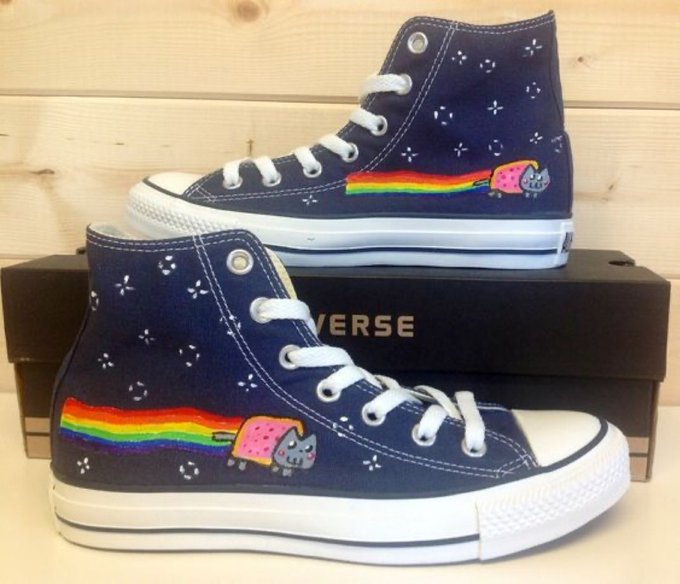 A pair of navy blue high-top Converse sneakers decorated with embroidered Nyan Cat rainbows, stars, and planets, sitting in front of a black Converse shoebox on a wooden surface.