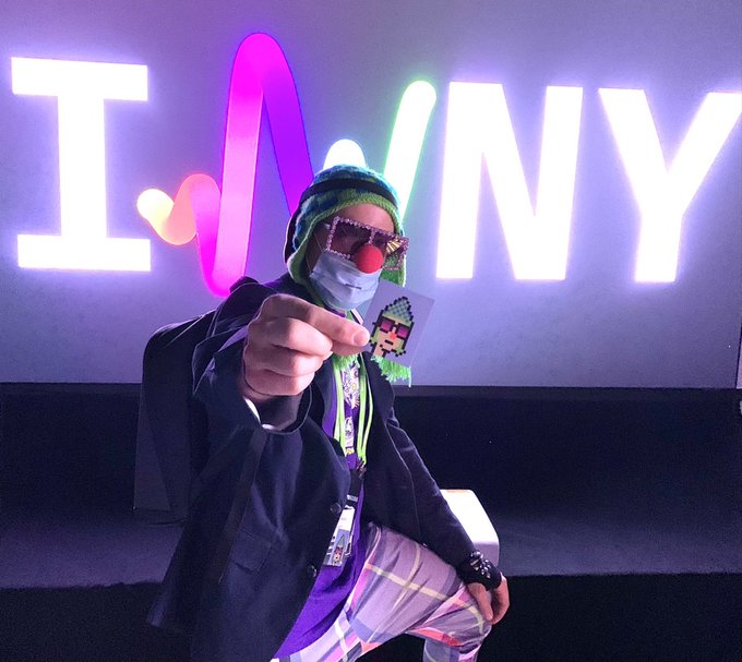 A person dressed as a clown with a red nose, mask, sunglasses, and striped beanie pointing at the camera, with a neon 'I ❤️ NY' sign in the background.