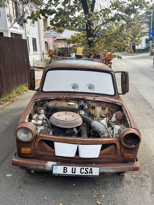A rusty, weathered car customized to resemble the character Tow Mater from the movie Cars, parked on a street with eyes painted on the windshield and a license plate reading 'B U CSA'.
