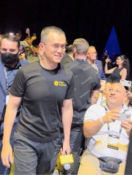 A smiling bald man in a dark t-shirt is captured in a candid photo at what appears to be a public event, surrounded by other people including someone holding a water bottle. The image relates to the post celebrating CZ (Changpeng Zhao, founder of Binance), highlighting his strong physique and bullish spirit despite his age, with the text suggesting his secret to staying fit is daily push-ups.