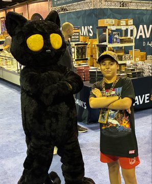 A photo taken at what appears to be a convention, showing a young boy in a graphic t-shirt and red shorts standing next to a person in a large black cat mascot costume.