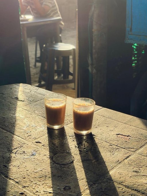 Two small glasses of brown liquid sit on a wooden table in a beam of sunlight, with a stool and person visible in the background.