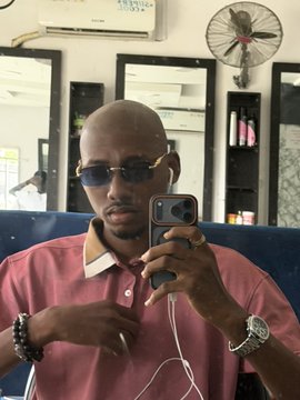 A bald man wearing sunglasses and a pink polo shirt takes a mirror selfie in a barbershop with a wall-mounted fan in the background, showcasing his freshly shaved head as part of a challenge to earn airdrops from @blknoiz06 and @lukebelmar.