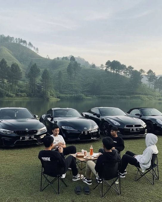 A group of young people sit in camping chairs around a small table near a serene lake, parked in front of four luxury sports cars (including a Toyota Supra, Subaru BRZ, Nissan GT-R, and Porsche). The license plates display "D 7371 VBJ", "D 1102 R", "D 1888", and "B 1945 GK", with the captioned post humorously suggesting this is what crypto holders' lives will look like in 2027.