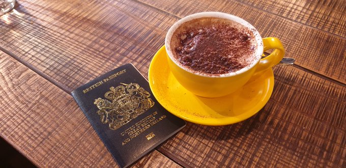 A yellow cup of hot chocolate on a saucer sits on a wooden table next to a British passport.