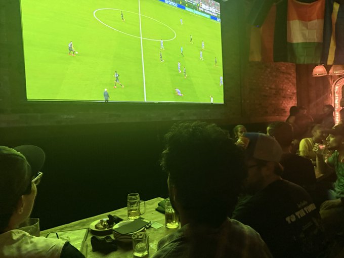The image shows a group of people sitting around a table, watching a soccer game on television. There are at least ten people visible in the scene, with some seated closer to the TV screen while others are further away. The TV screen displays a soccer match between two teams playing against each other. In addition to the spectators, there is also an assortment of drinks and food items spread across the table, indicating that they might be enjoying refreshments during the game.