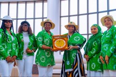 A group of women dressed in matching green jackets and hats posing together, with one holding what appears to be a golden plate or award.