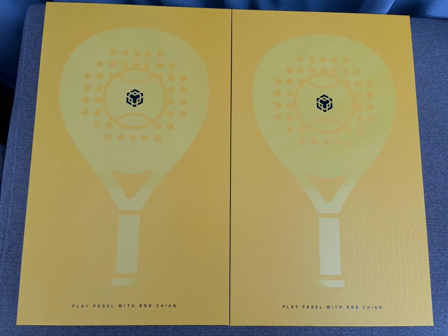 Two yellow promotional posters or boxes side by side featuring a stylized graphic of a padel racket, with text reading 'PLAY PADEL WITH BNB CHIAN' and a small cube logo.