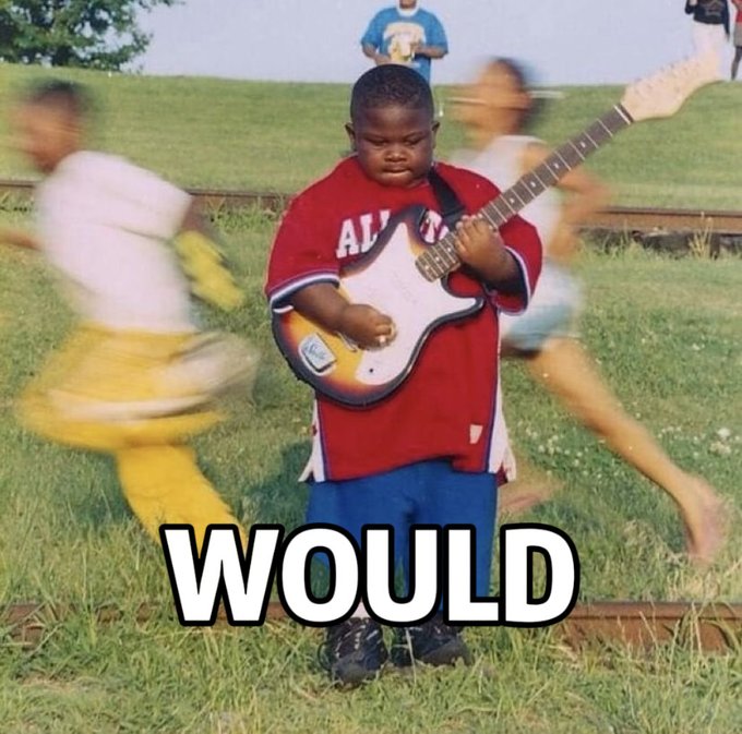 A young boy in an oversized red "ALL" jersey plays an electric guitar while blurred children run around him in a grassy field, with the word "WOULD" displayed at the bottom — humorously capturing the meme's theme that others may be active or chaotic, but he remains focused on rocking out.