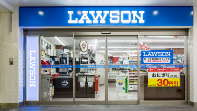 The exterior storefront of a LAWSON convenience store in Japan, featuring the iconic blue signage, glass entrance doors, and promotional posters advertising 30円 off on おにぎり (rice balls) and an open sale starting November 1st.